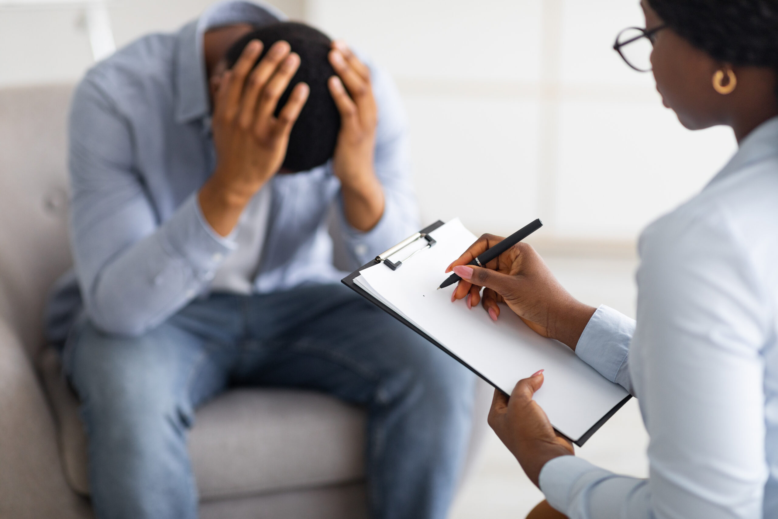 Psychotherapy concept. Depressed black guy holding his head at counselor's office, seeking professional help with mental disorder. Professional psychologist having session with desperate male patient